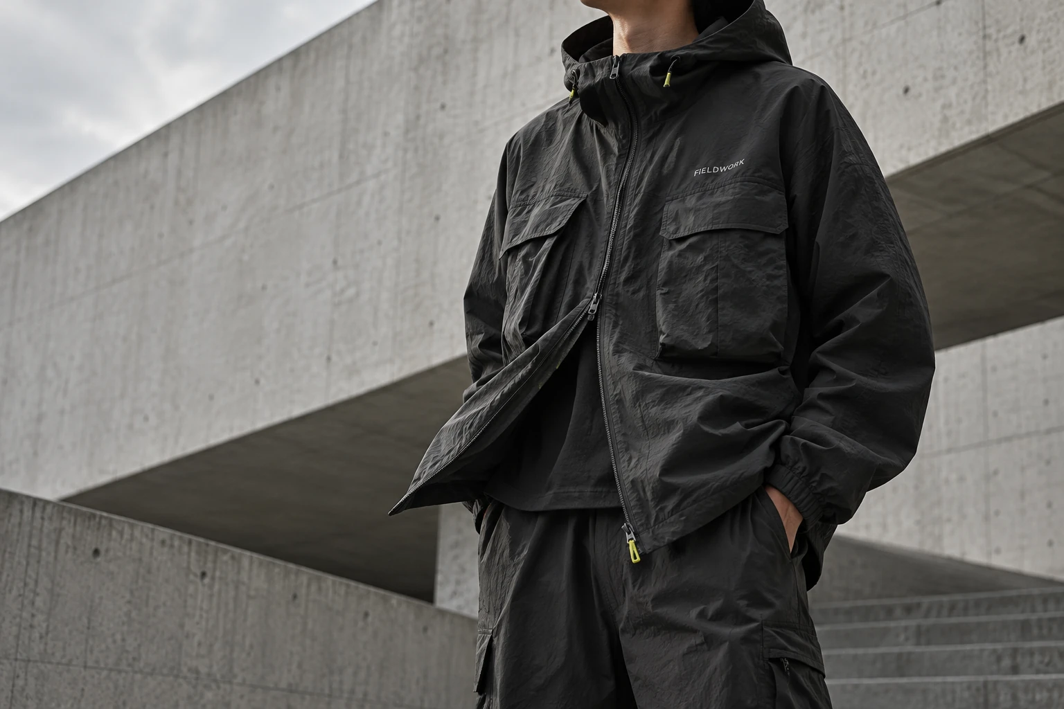 Editorial photograph of a fictional FIELDWORK charcoal shell worn against concrete architecture.